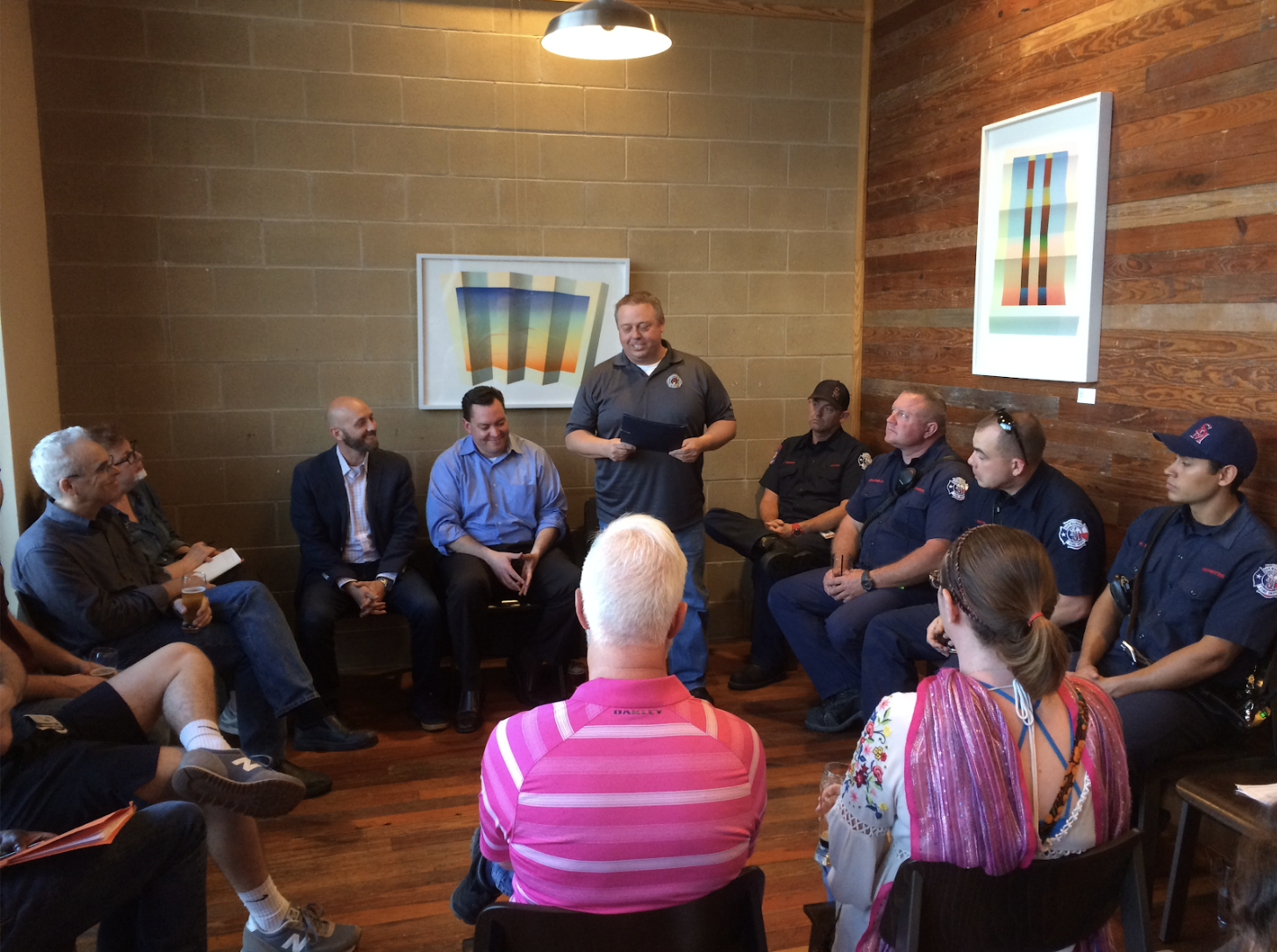 A group of about a dozen people in a crowded room, half of them in firefighter uniforms, sitting and listening attentively to a man standing.